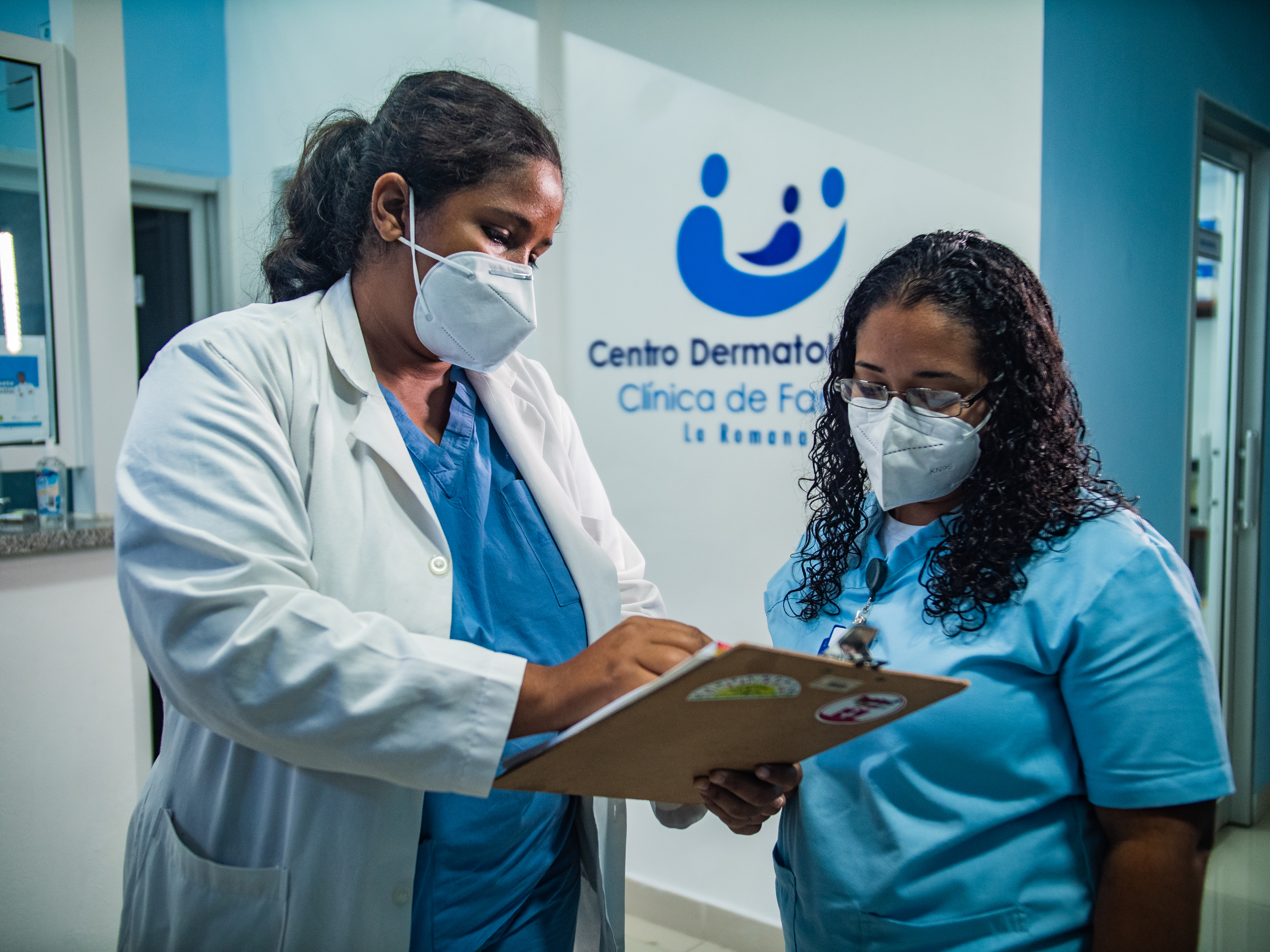Two female health providers at Clínica de Familia look at a clipboard together. One wears a lab coat and the other does not. They wear masks. 