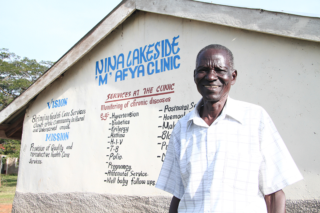 An middle-aged man stands in front of a health clinic in Kisumu, Kenya. The clinic has the name "Nina Lakeside 'M' Afya Clinic" on it.