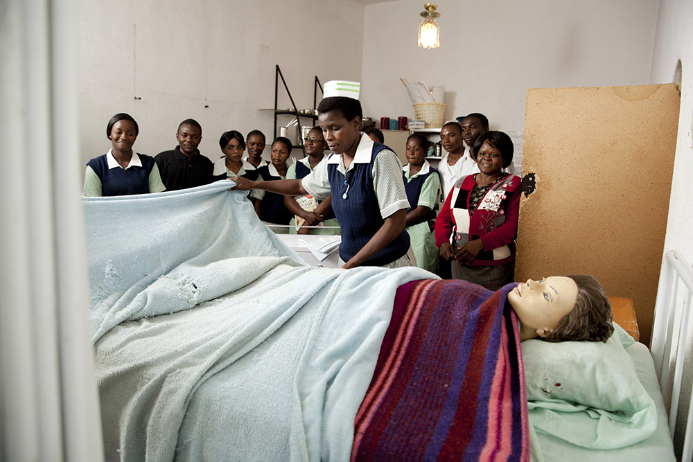 Nurses stand around a bed learning about patient care with a fake patient in the bed. 