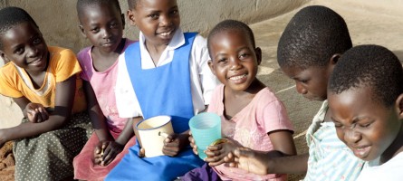 A group of African children sitting and smiling at the camera. Two of the children are holding cups in their hands. 
