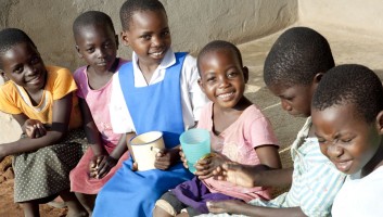 A group of African children sitting and smiling at the camera. Two of the children are holding cups in their hands. 
