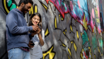 A man and a woman leaning against a wall covered in graffiti while looking at a phone